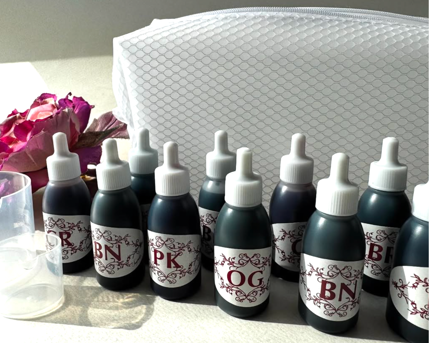 Set of ink bottles with labels on a surface with a white pouch and pink flowers in the background.
