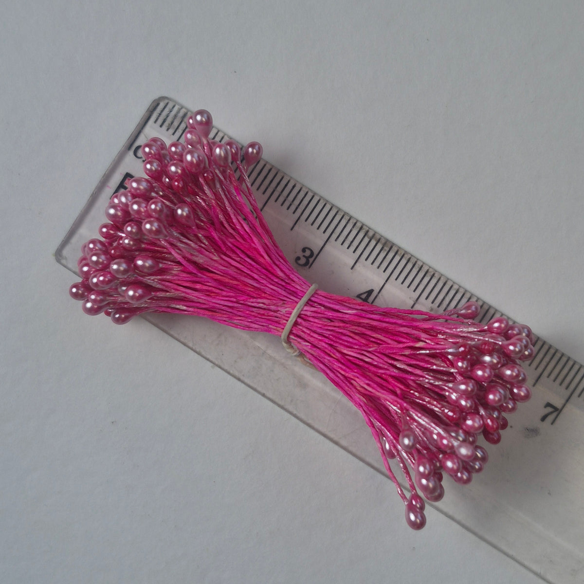 A bundle of size L Japanese miniature flower stamens, featuring bright pink pearlescent heads on vibrant raspberry cotton threads, arranged next to a transparent ruler